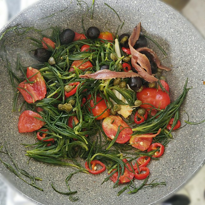 La barba dei frati viene saltata in padella con aglio, peperoncino, acciuga, capperi, olive, e pomodorini datterini.