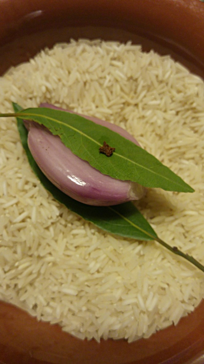 Una ciotola di riso basmati ancora da cuocere, assieme ad un piccolo scalogno isul quale sono state fissate due foglie di alloro tramite dei profumati chiodi di garofano.